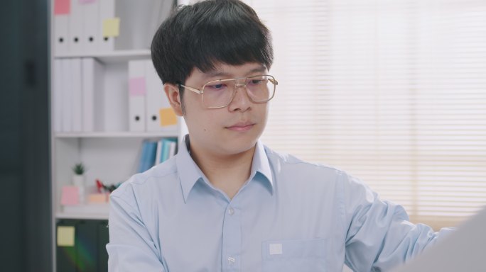 身穿蓝色正式衬衫的年轻亚洲商人坐在办公桌前，看着电脑打字，检查报告文书营销，以便在小办公室的会议板上