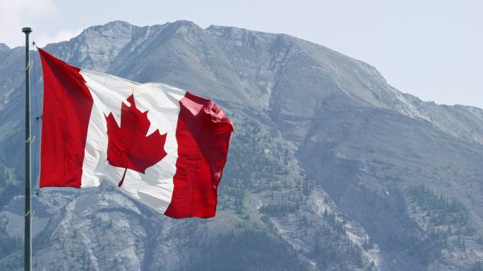 阳光明媚的一天，加拿大国旗随风飘扬，背景是一座山的慢镜头