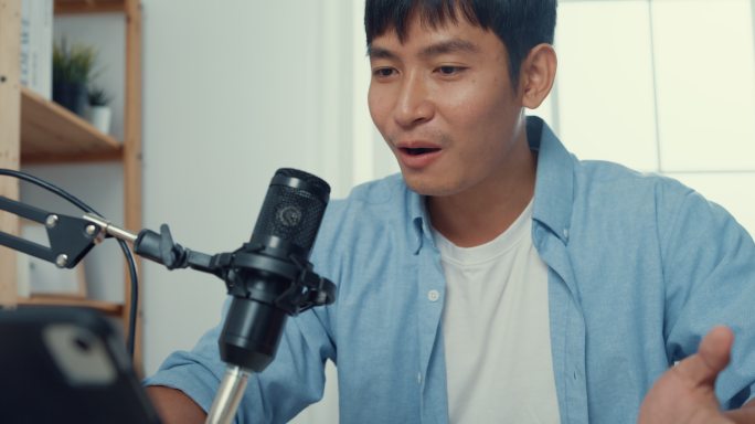 一位年轻的亚洲男子坐在家里客厅的办公桌上，用显微镜在笔记本电脑上录制播客。