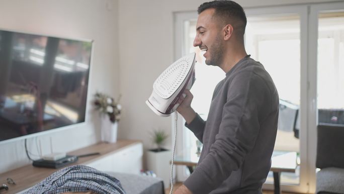 微笑的男人在家里整理衣服时用熨斗唱歌