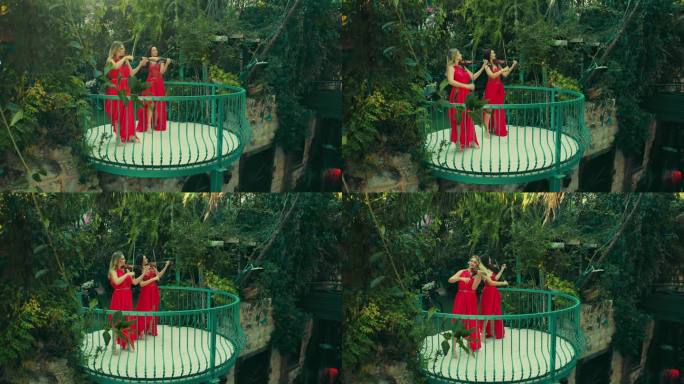 金发和黑发女小提琴手穿着红色连衣裙在树间的阳台上演奏小提琴