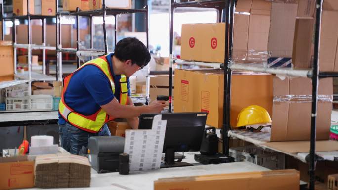 身穿制服的亚洲商人仓库工人在使用电脑检查发货时，在存储区扫描纸板箱