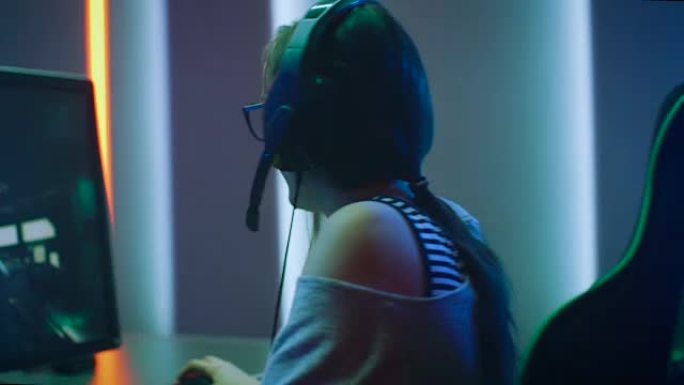 美丽的职业玩家女孩在她的个人电脑上玩在线FPS视频游戏，戴着眼镜和耳机的休闲可爱极客的弧光拍摄。霓虹