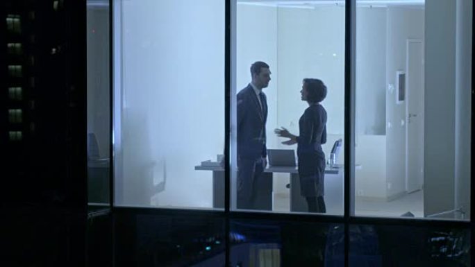 摩天大楼外的航拍镜头: 商人和女商人站在办公室窗户时见面并握手。晚上无人驾驶飞机拍摄商业区和在大城市