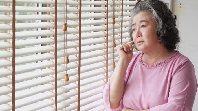 退休妇女独自在家。悲伤的老妇人独自站在窗户旁思考: 自然采光镜头中的悲伤和沮丧