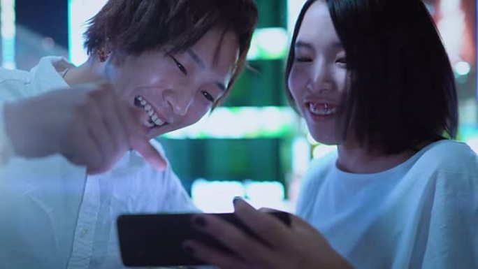 年轻的日本男孩和女孩夫妇聊天，大笑，使用手机和共享屏幕。在背景中模糊的广告广告牌和夜晚的城市灯光。