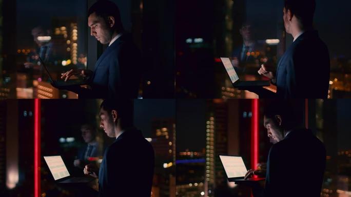 深夜在办公室里，成功的商人在工作时拿着笔记本电脑。在带有城市灯光的商业区窗口中。