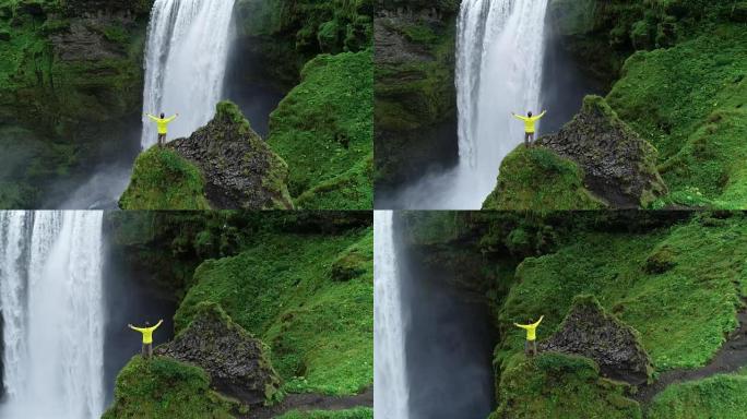 慢动作男子，双臂高举在悬崖顶上，站在瀑布前庆祝成功的旅行和冒险