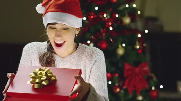 戴圣诞帽的女孩许愿并打开圣诞礼包。假期和新年的概念。这个女孩很开心，手里拿着圣诞礼物微笑着。