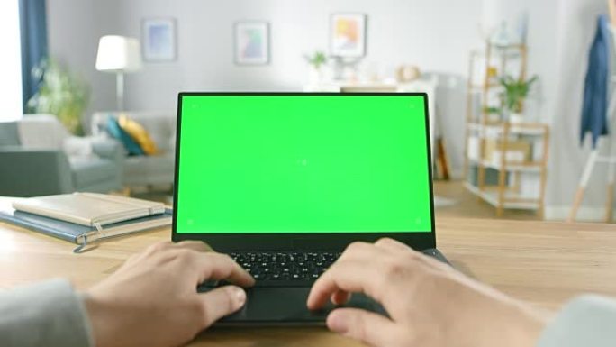 第一人称特写镜头男子坐在舒适的客厅的桌子旁，使用带有绿色模拟屏幕的笔记本电脑。