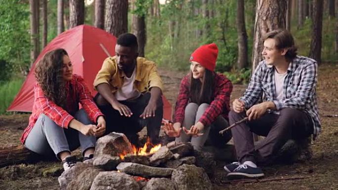 年轻的男女旅行者围坐在火炉旁，讲故事，大笑，帅哥在火焰中扔柴火。帐篷和背包是可见的。