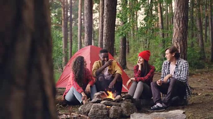 快乐的女孩和男孩徒步旅行者坐在营地附近的火旁聊天，分享故事并玩乐。绿树、现代帐篷和火焰可见。