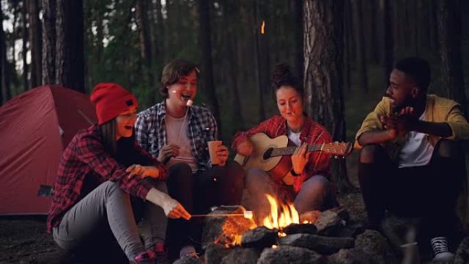 快乐的冒险徒步旅行者围坐在火炉旁，唱歌和弹吉他，在黑暗的夏日傍晚吃棉花糖和拍手。森林中的帐篷是可见的
