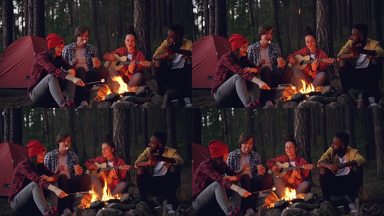 快乐的冒险徒步旅行者围坐在火炉旁，唱歌和弹吉他，在黑暗的夏日傍晚吃棉花糖和拍手。森林中的帐篷是可见的