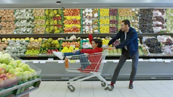 在超市: 男人推着购物车，女人坐在购物车里，幸福的夫妇在手推车里穿过商店的新鲜农产品区玩得很开心。慢