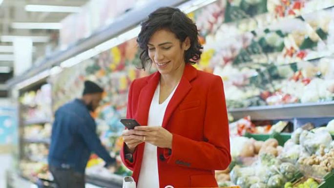 在超市: 美女站在商店的新鲜农产品区时使用智能手机。女人沉浸在网上冲浪在她的手机背景五颜六色的水果和