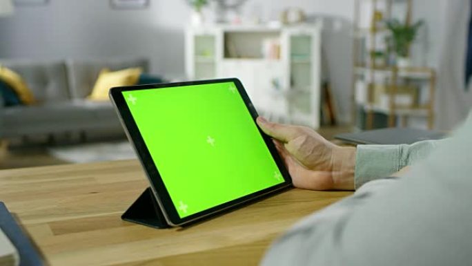 一名男子坐在办公桌前拿着并观看绿色模拟屏幕数字平板电脑的肩膀镜头。背景舒适的客厅。