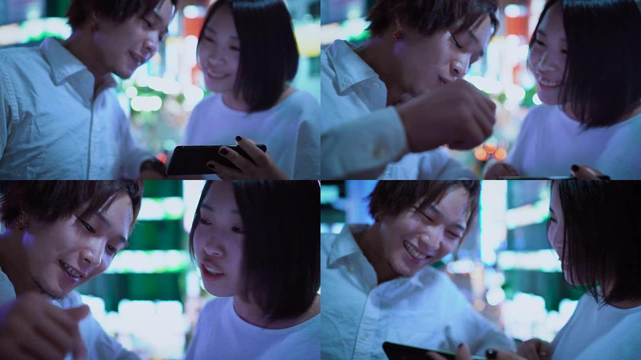 年轻的日本男孩和女孩夫妇聊天，大笑，使用手机和共享屏幕。在背景中模糊的广告广告牌和夜晚的城市灯光。