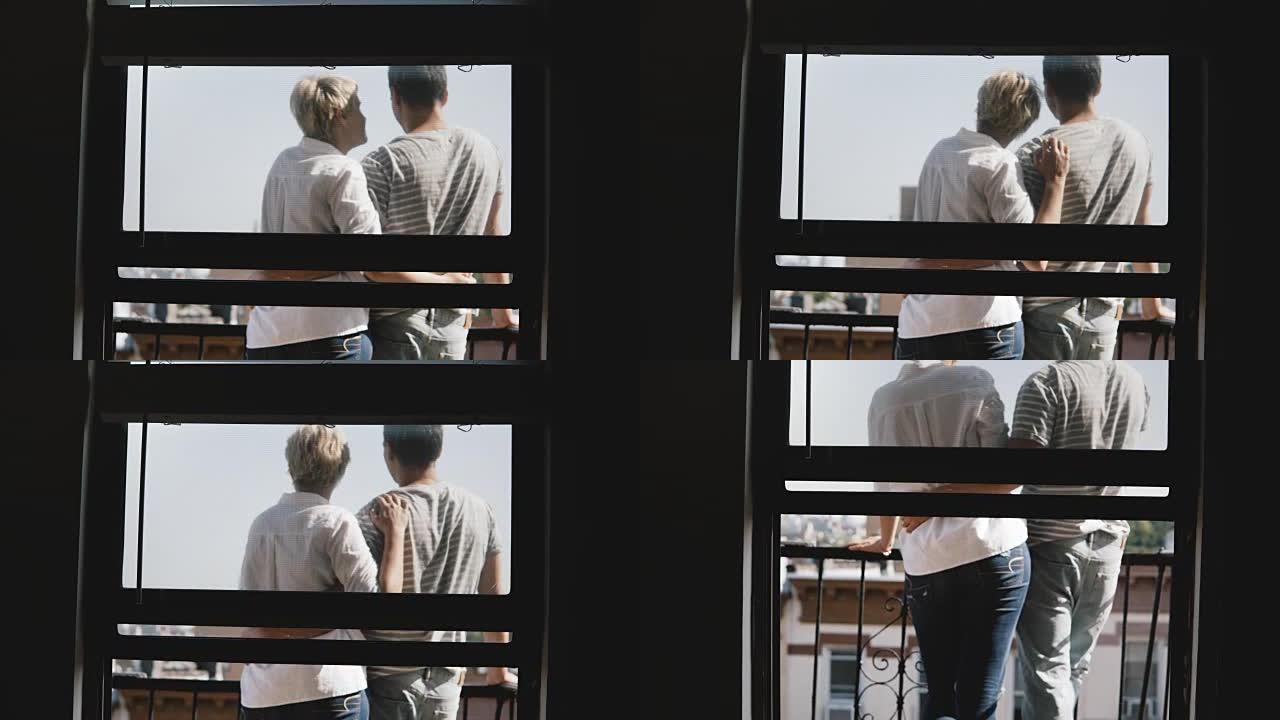 可爱的年轻浪漫情侣站在一个可爱的阳光明媚的小阳台上轻轻亲吻，透过公寓的窗户可以看到