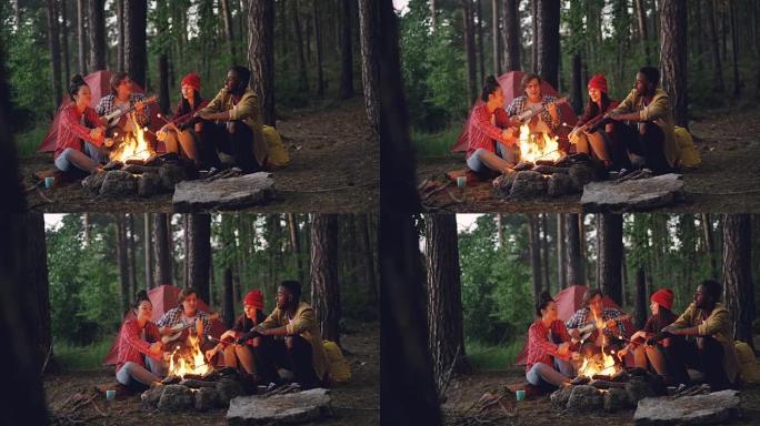 轻松的男女旅行者在森林的篝火旁唱歌，弹吉他和烤棉花糖。绿树和帐篷是可见的。