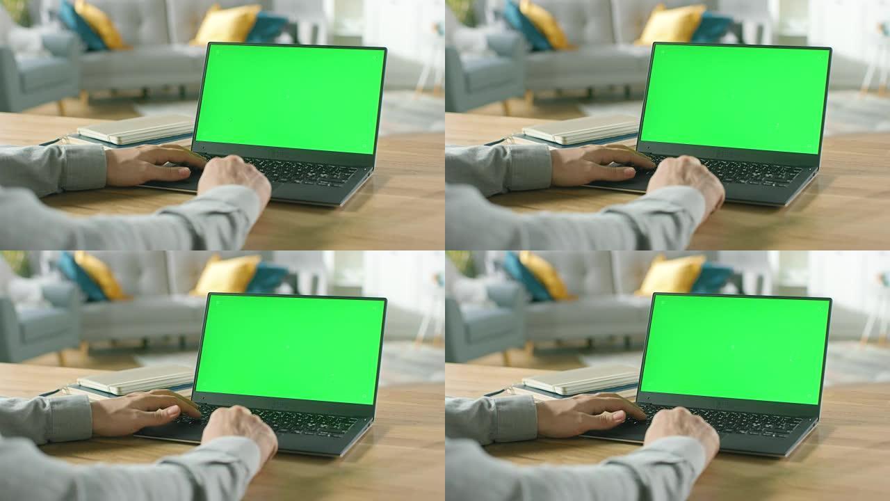 一个男人的特写镜头坐在他舒适的客厅的桌子旁，使用带有绿色模拟屏幕的笔记本电脑。