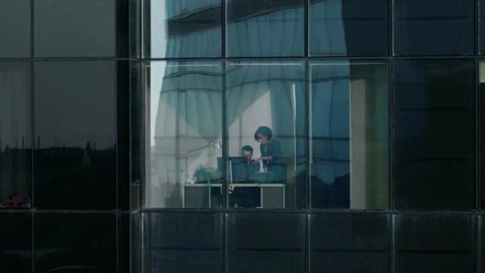 摩天大楼外的航拍画面: 商人和女商人坐在办公室窗户旁的办公桌旁谈论生意。金融区和在大城市工作的商人的