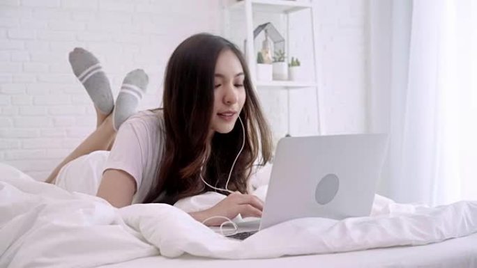 美丽的亚洲女人躺在卧室的床上玩电脑。快乐的女性在家中戴着耳机听音乐并在笔记本电脑中搜索歌曲。生活方式