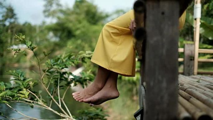 穿着黄色西装的亚洲女人放松，微笑，坐在木墩上，脚在水面上晃来晃去