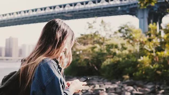 带背包的年轻旅行者女孩的侧视图，用智能手机慢动作沿着平静的布鲁克林大桥河岸行走
