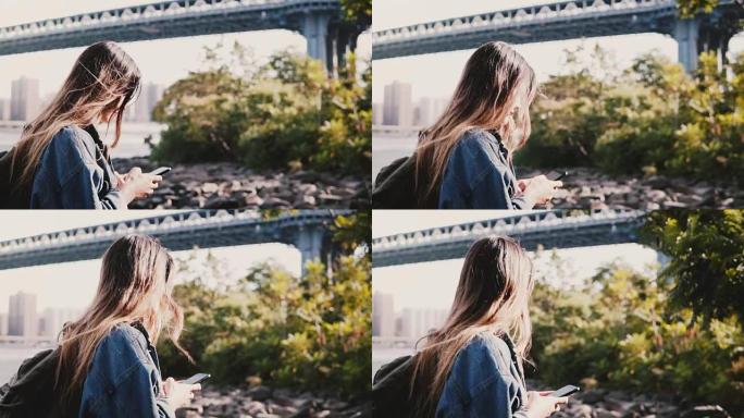 带背包的年轻旅行者女孩的侧视图，用智能手机慢动作沿着平静的布鲁克林大桥河岸行走