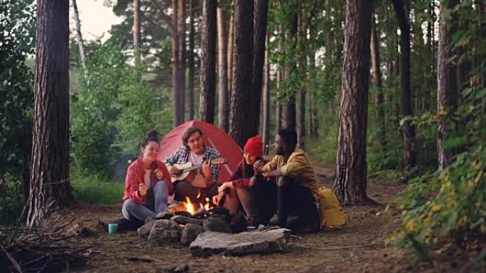 穿着休闲服的游客正在弹吉他，而他的朋友们则在秋天的篝火旁做饭和吃甜棉花糖。人和营地概念。