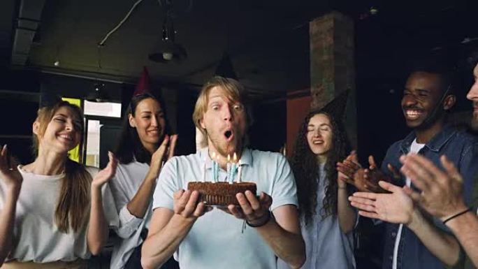 脸庞富有表情的大胡子家伙正在许愿，并在生日蛋糕上吹蜡烛，而他的同事们正在鼓掌祝贺他。
