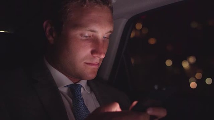 年轻的商人晚上在繁忙的城市的汽车后座上用手机发短信