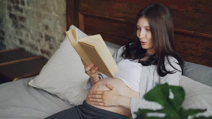 微笑的女孩期待着母亲向她未出生的婴儿大声朗读书，微笑着在现代公寓的床上休息，抚摸着她的腹部。文学与人