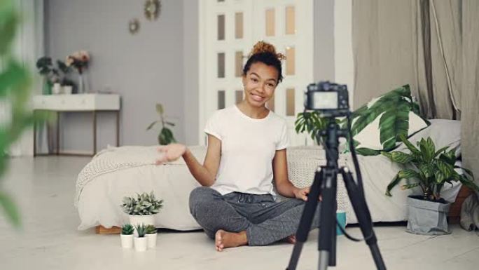 好看的非洲裔美国女孩创意博客作者正在录制有关坐在她公寓地板上的植物并在三脚架上看着相机的视频。