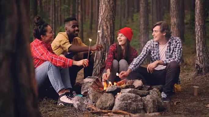 饥饿的旅行者在火上烹饪棉花糖，并在篝火旁交谈时用木棒吃，人们在享受甜食时会说话和大笑。