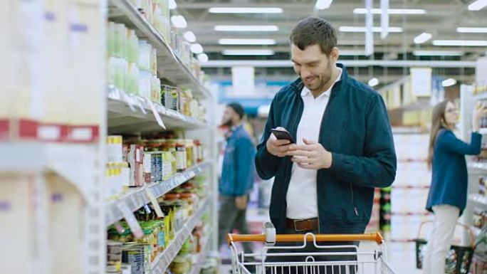 在超市: 英俊的男人使用智能手机，走过罐头货架。他正推着购物车穿过罐头食品区。