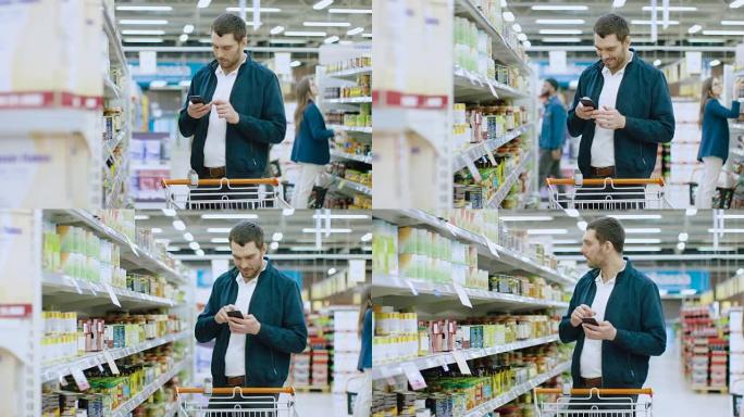 在超市: 英俊的男人使用智能手机，走过罐头货架。他正推着购物车穿过罐头食品区。