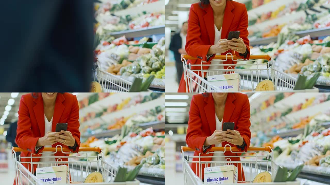 在超市: 女人使用智能手机，靠在商店新鲜农产品区的购物车上。在大商场里，女人在手机上浏览互联网。慢动
