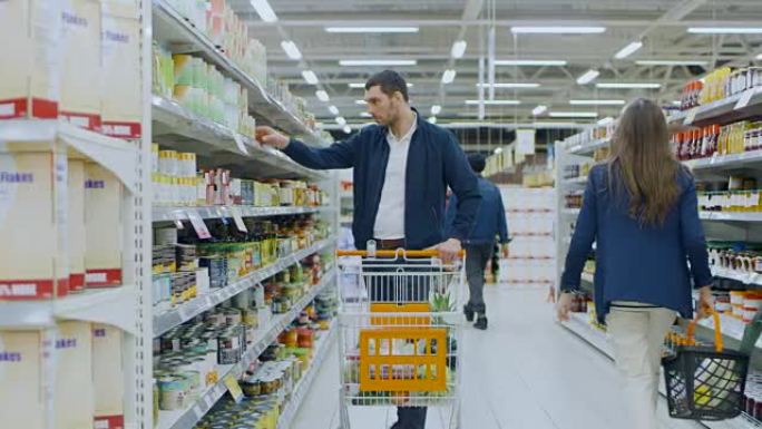 在超市: 英俊的男人在罐头食品的货架上浏览，看着锡罐，但决定不买。他继续带着购物车穿过商店的不同部分
