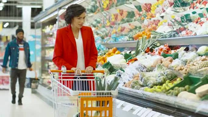 在超市: 带购物车的美丽年轻女子在新鲜农产品区选择蔬菜，将它们放入手推车。