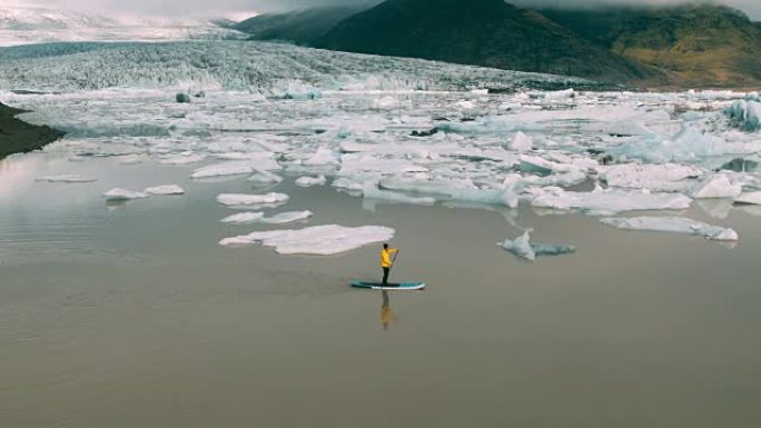 在冰岛的冰川泻湖中，有巨大冰山的男子划桨站立桨板的空中射击