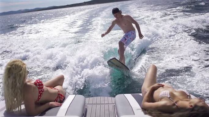 年轻人在船后冲浪，两个女孩在船上晒日光浴