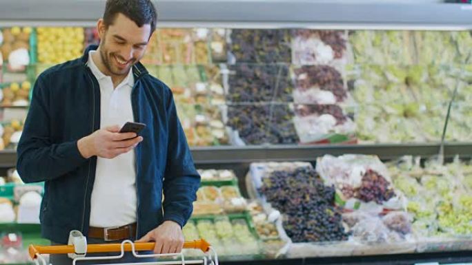 在超市: 英俊的男人站在商店的新鲜农产品区时使用智能手机。一名男子沉浸在网上冲浪，在他的手机背景五颜