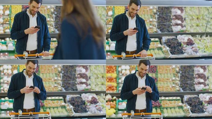 在超市: 英俊的男人站在商店的新鲜农产品区时使用智能手机。一名男子沉浸在网上冲浪，在他的手机背景五颜