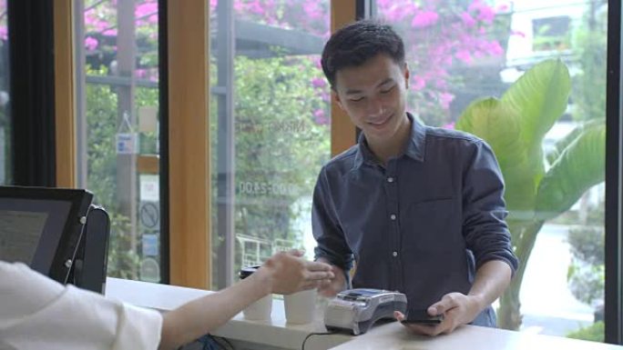 在咖啡馆里，美丽的亚洲女人为通过非接触式手机向信用卡系统付款的顾客制作外卖咖啡