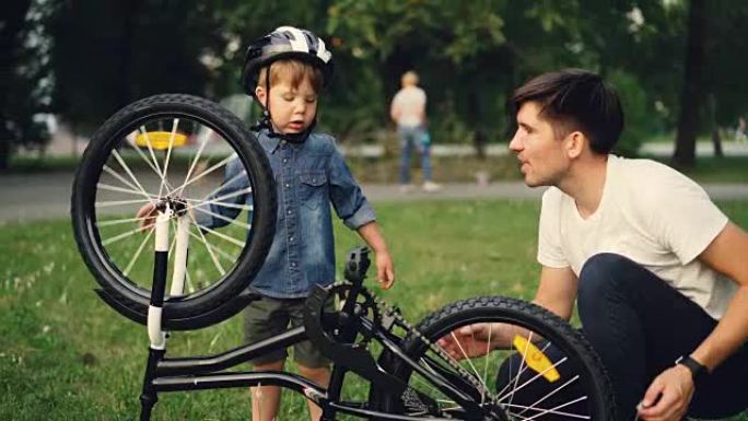 夏天，当他的父亲在公园的草坪上与他交谈时，小男孩正在旋转自行车车轮和踏板。家庭、休闲和积极的生活方式