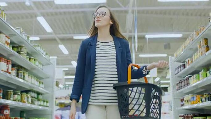 在超级市场:美丽的年轻女子拿着购物篮走过罐头区，浏览。有很多通道的大商店。