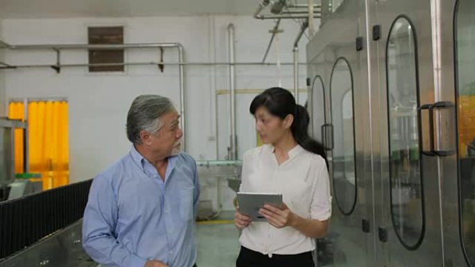 一位成熟的男性工厂经理在一位年轻的女性协助下，看着平板电脑，并在背景中讨论瓶子生产线的质量问题