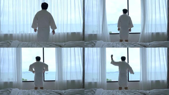4k镜头亚洲男子走路打开窗户窗帘，早上在豪华酒店卧室醒来时伸展自己的场景，生活方式和休闲概念